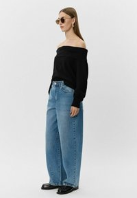 Melnā plecu atklātā džemperis, gaiši zilas platās bikses un melnas kurpes. Aksesuāri ietver apaļas saulesbrilles un izteiksmīgas auskarus.