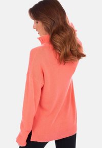 Pull en corail surdimensionné avec un col haut, un ourlet côtelé et un tricot texturé. Porté sur des leggings noirs, affichant une coupe et une silhouette décontractées.