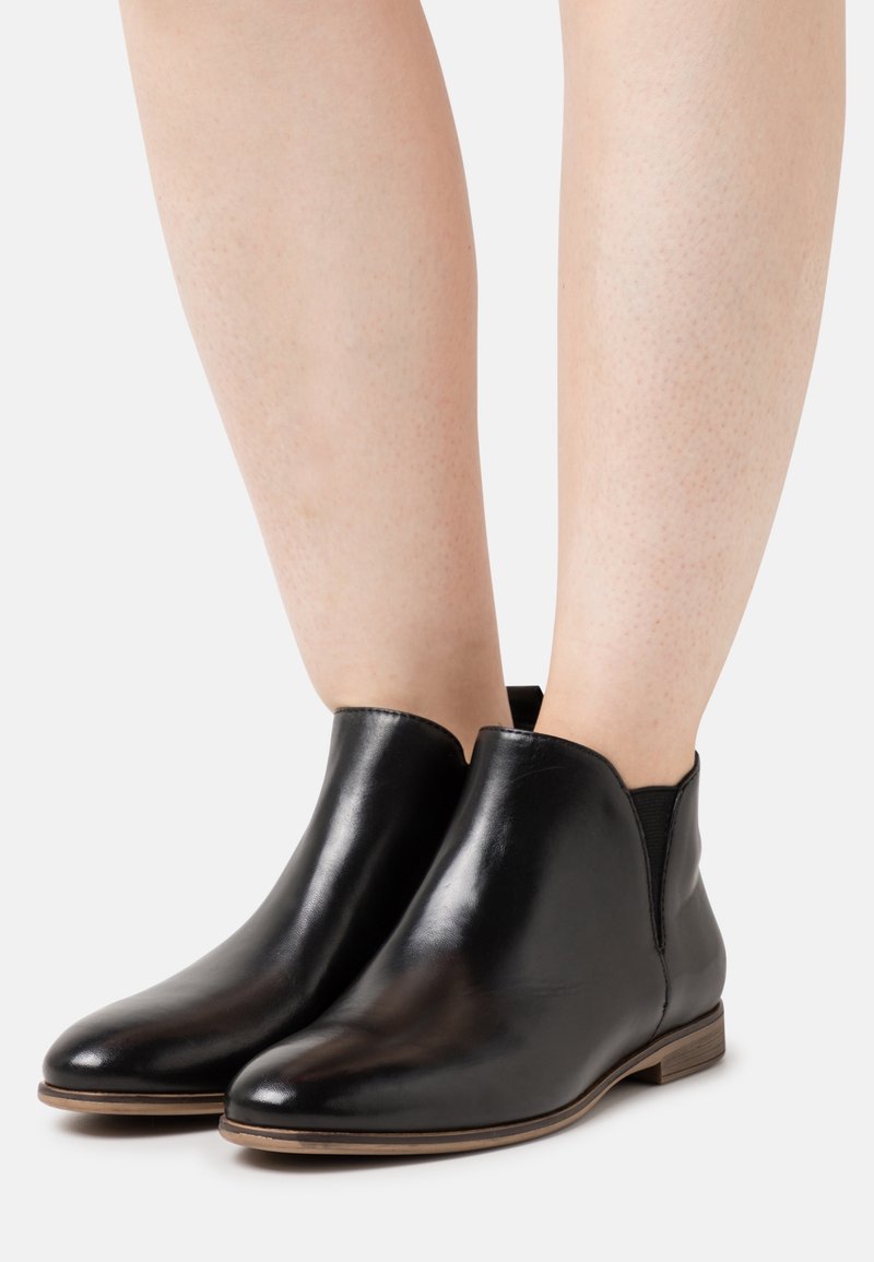 Bottes basses en cuir noir avec talons en bois bas portées sur des jambes nues sur un fond clair uni.