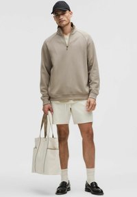 Beige zip-neksweater, crèmekleurige shorts, zwarte pet, met een beige totebag met tan accenten. Geweven stof, relaxte pasvorm en contrasterende kleuren.
