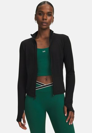 Vrouw draagt een zwart jack met rits over een groene sportbeha en groene legging met witte en zwarte kruisstrepen bij de taille.