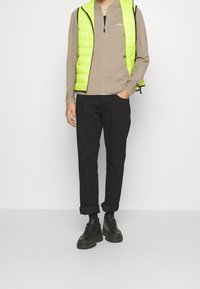 Neonově zelená prošívaná vesta přes béžový top na zip, doplněná o černé kalhoty s ohrnutými lemy a černé šněrovací boty. Jednoduchý design a materiály.