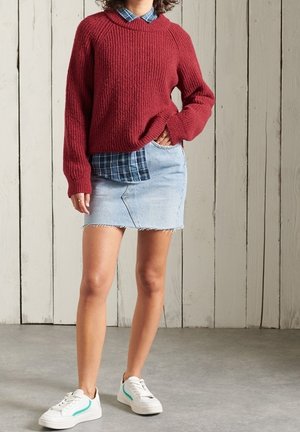 Personne portant un pull en tricot rouge par-dessus une chemise à carreaux bleue, une jupe en denim clair et des baskets blanches, debout contre un mur en planches de bois.