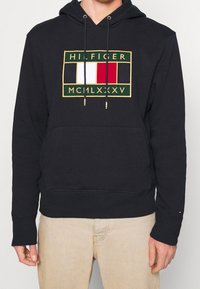 Felpa blu navy con cordoncini, caratterizzata da un logo ricamato in verde, bianco, rosso e dettagli dorati. Tessuto morbido e texturizzato.