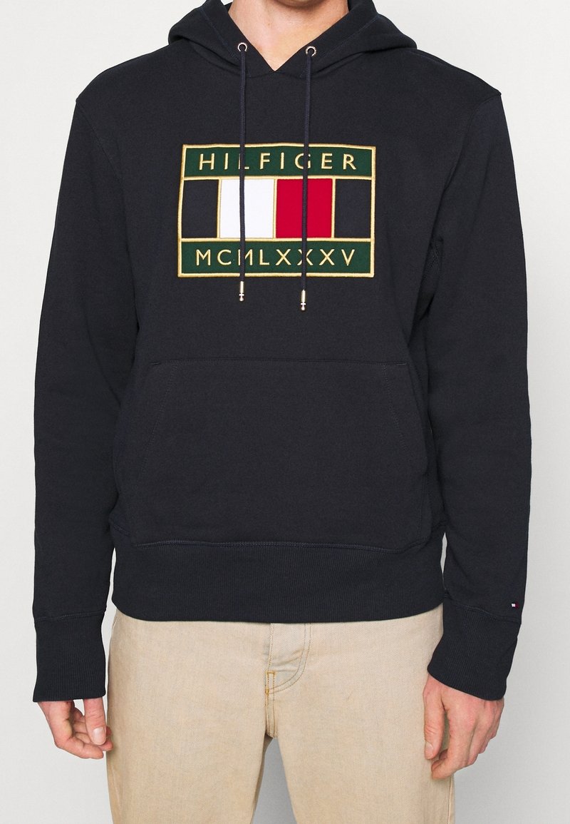 Felpa blu navy con cordoncini, caratterizzata da un logo ricamato in verde, bianco, rosso e dettagli dorati. Tessuto morbido e texturizzato.