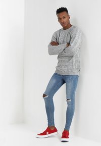 Suéter gris de punto trenzado, jeans azul ajustados con rodillas desgastadas y zapatillas rojas con acentos blancos. Modelo posando con los brazos cruzados.