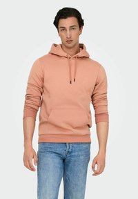Persikofärgad hoodie i bomullsblandning, med en framficka, dragsko och ribbade muddar. Bärs med blå jeans.