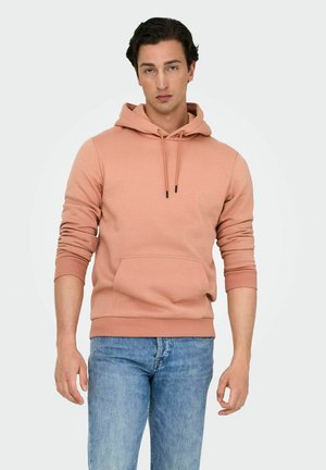 Peachfarvet hættetrøje lavet af bomuldsblanding, med en frontlomme, snøre i hætten og ribbede manchetter. Båret med blå jeans.