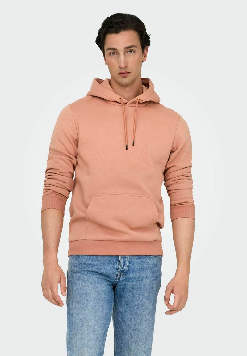 Persikofärgad hoodie i bomullsblandning, med en framficka, dragsko och ribbade muddar. Bärs med blå jeans.