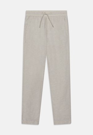 Person iført lysegrå bukser med den ene hånd i lommen og hvide sneakers står mod en ensfarvet baggrund.