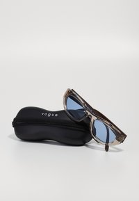 Óculos de sol com uma armação cinza translúcida e lentes com tom azulado, exibidos ao lado de um estojo preto com fecho éclair e o logótipo "vogue".