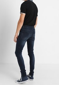 Persona con jeans ajustados azul oscuro, camiseta de polo negra de manga corta con ribete blanco y zapatillas altas azul marino, de pie de lado.