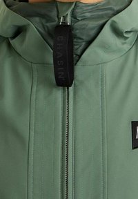 Green zip-up jacket featuring a high collar, textured fabric with vertical stitching, and a black pull tab with logo detail.