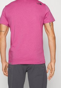 Magenta tričko s krátkým rukávem vyrobené z bavlny, s malým černým logem na horní části zad, přímým lemem a kulatým výstřihem.