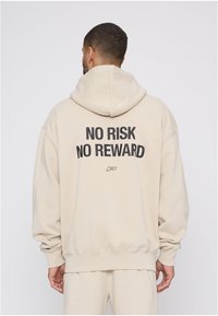 Bež dukserica s kapuljačom, opuštenog kroja, s rebrastim manžetnama i rubom. Na stražnjoj strani crni tekst: "NEMA RIZIKA NEMA NAGRADE." Glatka tekstura.