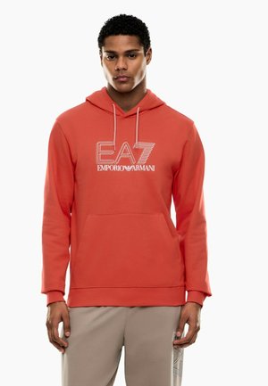 Ung mand iført orange EA7 Emporio Armani hættetrøje og beige bukser, står vendt fremad med neutralt ansigtsudtryk.