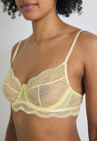 Yellow lace bra featuring sheer lace cups, scalloped edges, and thin straps. Detailed lace pattern enhances visual texture.