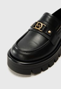 Mocassin en cuir noir avec semelle épaisse à rainures et boucle logo MK dorée sur la bride supérieure.