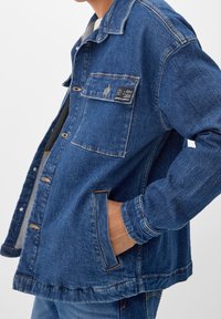 Chaqueta de mezclilla en azul medio con un corte relajado, que cuenta con un cierre a presión, bolsillo en el pecho y detalles de costura sutiles.