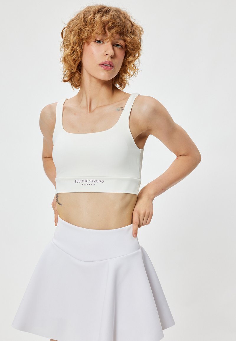 Top corto atlético blanco con escote cuadrado, diseño ajustado y detalle del logo en el dobladillo, combinado con una falda blanca de corte evasé.