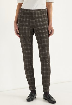 Kvinde iført tætsiddende brune og grå ternede bukser kombineret med sorte loafers og en hvid bluse, stående mod en ensfarvet baggrund.