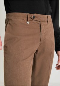 Pantalon marron à motif avec un tissu texturé, doté d'une fermeture à bouton, de passants pour ceinture et de poches latérales, présenté sur un mannequin portant une chemise noire.