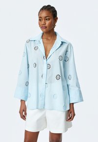 Hellblaue Button-Down-Hemd mit floralem Ausschnittmuster, langen Ärmeln und klassischem Kragen. Getragen mit weißen Shorts.