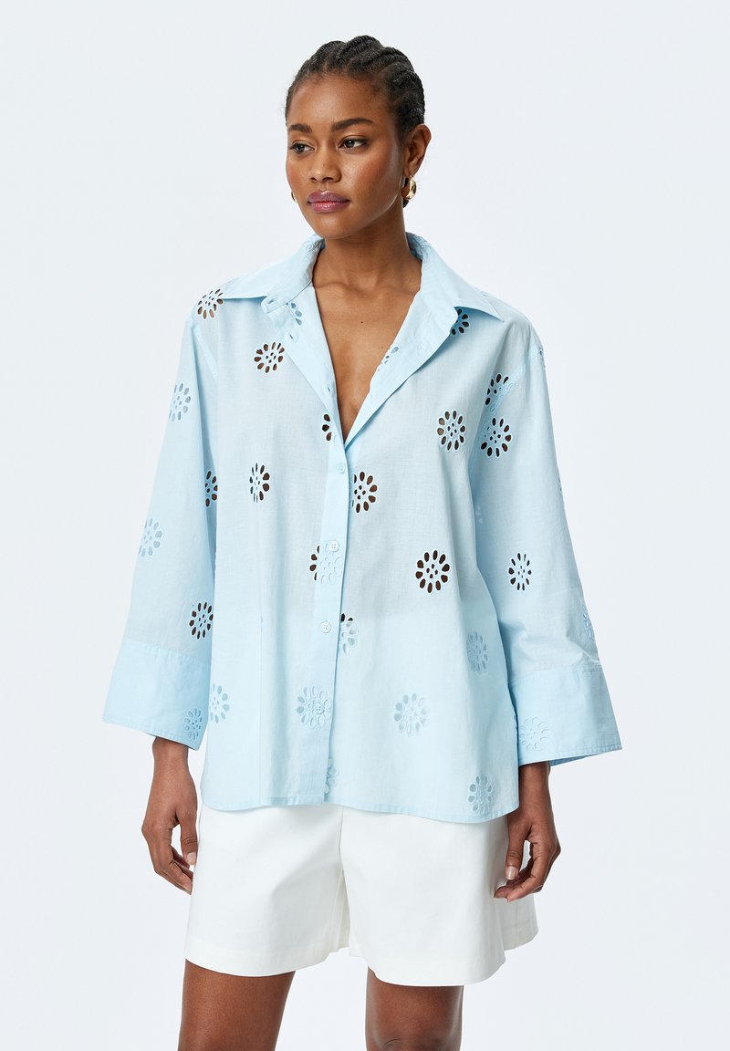 Hellblaue Button-Down-Hemd mit floralem Ausschnittmuster, langen Ärmeln und klassischem Kragen. Getragen mit weißen Shorts.