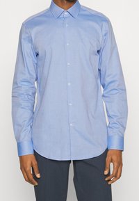 Světlomodrá košile z hladké látky, s límečkem, dlouhými rukávy a zaobleným lemem s jemnými knoflíkovými detaily.