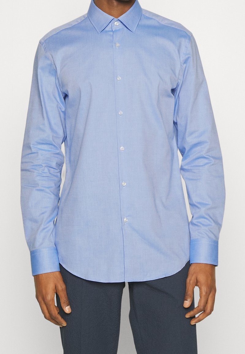 Světlomodrá košile z hladké látky, s límečkem, dlouhými rukávy a zaobleným lemem s jemnými knoflíkovými detaily.