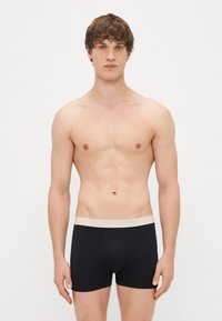 Črne bombažne bokserice z bež elastičnim pasom, ki imajo prilegan dizajn in gladko teksturo. Brez vidnih vzorcev ali akcentov.