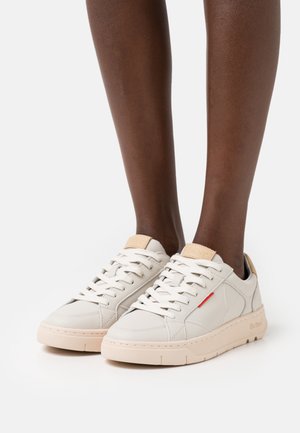 Zapatillas ligeras de cuero crema con acentos beige, con una textura suave, punta redonda y cierre de cordones con un sutil detalle en rojo.