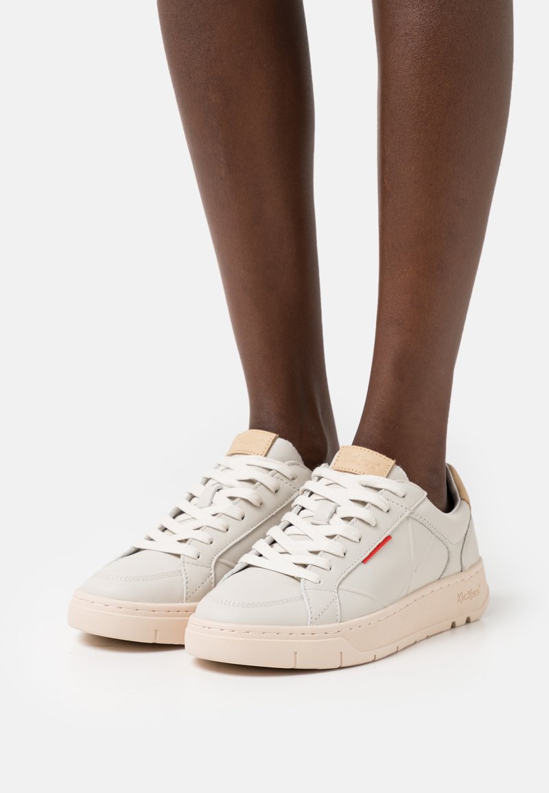 Sneaker leggere in pelle crema con accenti beige, caratterizzate da una superficie liscia, punta tonda e chiusura con lacci con un sottile dettaglio rosso.