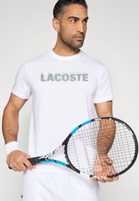 Biała koszulka z krótkim rękawem z zielonym logo "LACOSTE"; zawodnik trzyma czarno-niebieską rakietę tenisową z teksturowanym uchwytem.