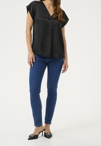 Haut court en satin noir à manches courtes avec un col en V, associé à un jean skinny bleu et des talons noirs pointus ornés de boucles décoratives.