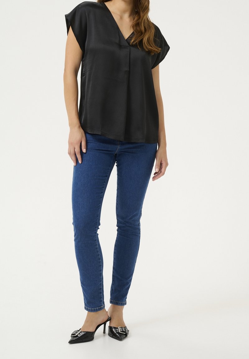 Haut court en satin noir à manches courtes avec un col en V, associé à un jean skinny bleu et des talons noirs pointus ornés de boucles décoratives.