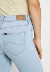 Pantalones vaqueros de mezclilla azul claro con un ajuste ceñido, que cuentan con una etiqueta de cuero con "Lee" en la cinturilla y costuras en contraste en los bolsillos.