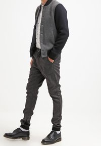 Grijs en zwarte varsityjack met ribgebreide cuffs, een witte blouse met stippen, donkergrijze tapered sweatpants en zwarte veterschoenen.