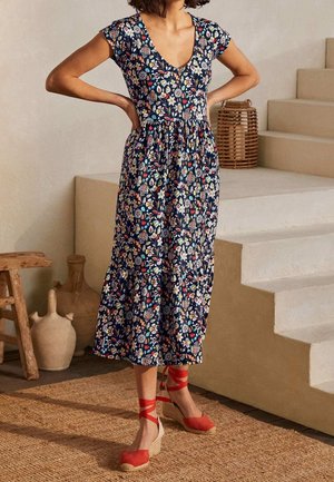 Femme debout, mains sur les hanches, portant une robe midi fleurie bleu marine et des sandales compensées en espadrille rouges, sur un tapis tissé près des escaliers.