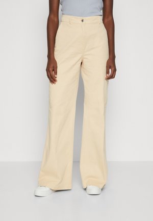 Personne portant un pantalon beige à jambes larges et taille haute avec des baskets blanches, debout devant un fond clair uni.