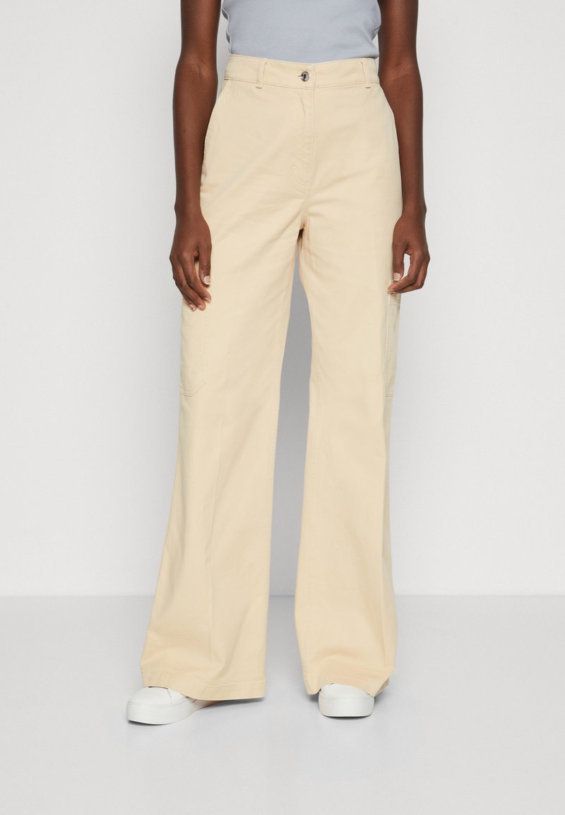 Personne portant un pantalon beige à jambes larges et taille haute avec des baskets blanches, debout devant un fond clair uni.