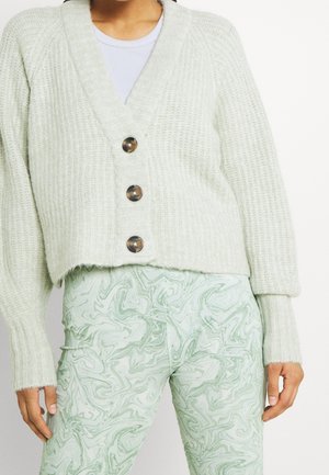 Une personne porte un cardigan crème à la texture travaillée et à trois boutons, sur un haut blanc et un pantalon à motifs vert pâle.