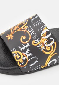 Sandale noire à glissière avec une large bride ornée de motifs jaunes et blancs élaborés sur un fond texturé. Comprend le lettrage de la marque.