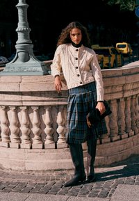 Kremowy kardigan w splot kablowy z czarnymi guzikami, plisowana spódnica midi w niebiesko-zieloną kratę, brązowe kozaki do kolan i czarna kopertówka.