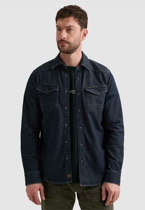Homme aux cheveux courts et bruns avec une barbe, portant une chemise en jean sombre boutonnée sur un t-shirt noir et un pantalon vert olive, debout devant un fond uni.