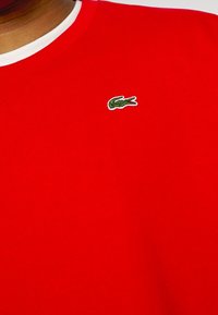 Czerwona bawełniana bluza z białym dekoltem w kształcie łódki, z małym zielonym haftowanym logo krokodyla po lewej stronie klatki piersiowej.