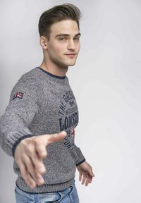 Pullover grigio lavorato a maglia con orlo e polsini a coste, dotato di un logo e di una toppa con la Union Jack sulla manica. Indossato con jeans blu.