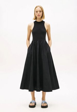 Mujer con cabello rubio corto, vestida con un vestido midi negro sin mangas con bolsillos y sandalias azul claro, posando sobre un fondo blanco.