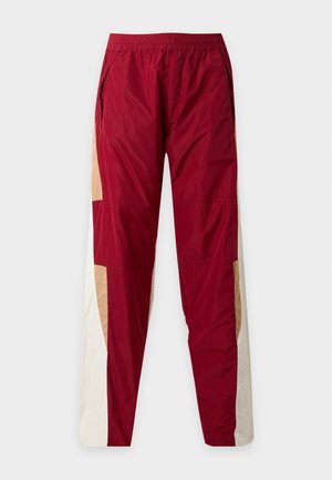 Hombre que lleva una camiseta blanca Dsquared2, pantalones deportivos rojos y beige, zapatos marrones, gorra negra Dsquared2 y dos collares de cruz dorados, de pie con las manos en los bolsillos.