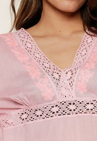 Różowa bluzka z misternymi koronkowymi detalami na dekolcie i klatce piersiowej, z kwiatowymi wzorami oraz lekkim, strukturalnym materiałem.